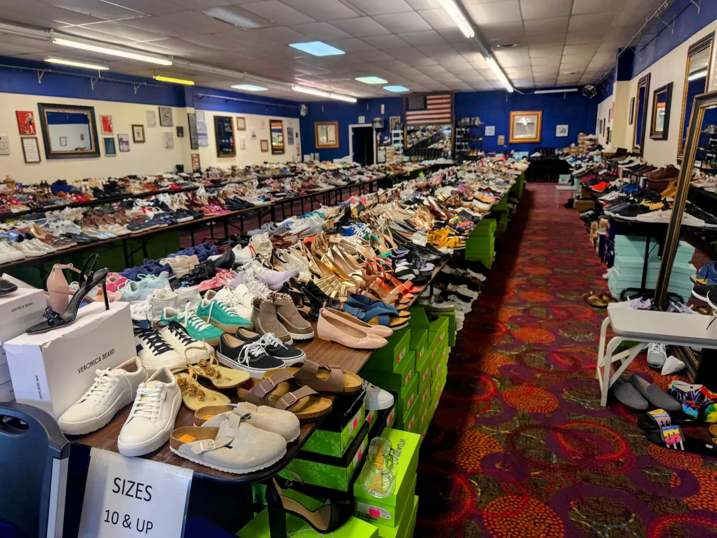 A table of shoes at The Shoe Room with stacked designer boxes — Veronica Beard, Sam Edelman — beside sneakers, Birkenstocks, and sandals.
