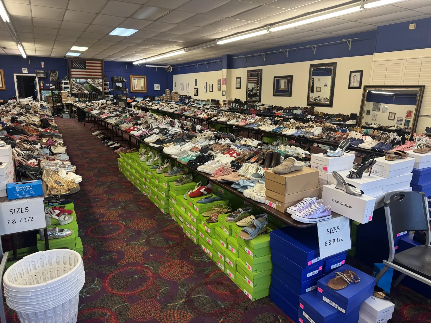 The main aisle of The Shoe Room lined with long tables of shoes sorted by size, boxes stacked underneath, and a basket waiting by the front.